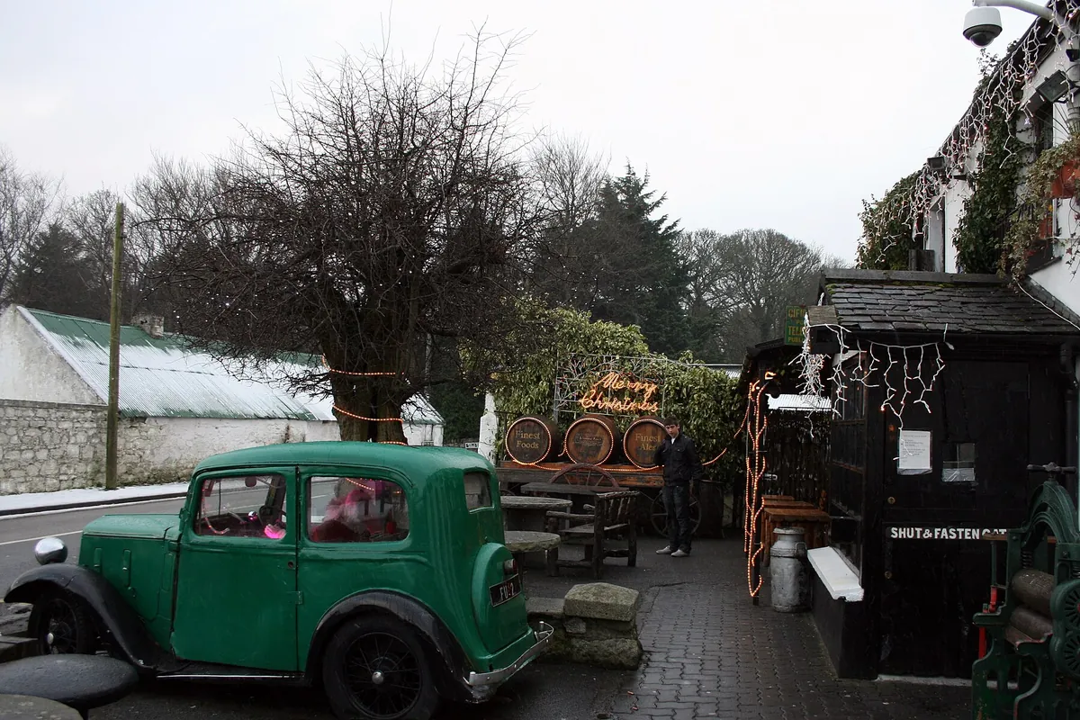 Une voiture verte vintage et des tonneaux en bois se dressent sur une cour pavée à côté du porche en bois sombre d'un pub, avec des arbres dénudés et une dépendance blanche en arrière-plan