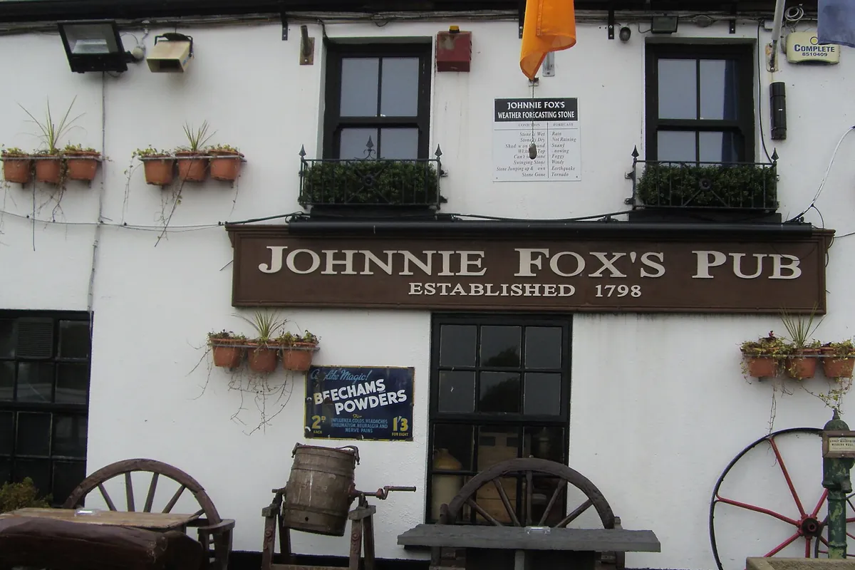 Weiß getünchte Wand eines Pubs mit einem braunen Schild mit der Aufschrift Johnnie Fox's Pub, Established 1798, Blumentöpfe an der Wand sowie alte Wagenräder und ein Fass davor
