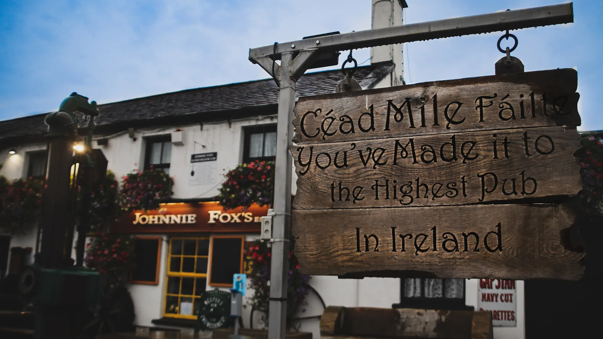 Et udskåret træskilt hænger fra en jernstang i skumringen med teksten Céad Mile Fáilte, You've made it to the highest pub in Ireland, foran den hvide pub med det lysende orange Johnnie Fox's-skilt